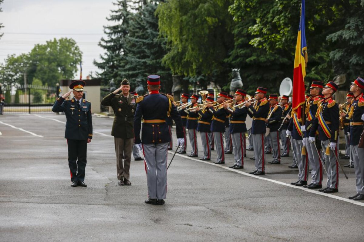 Two uniformed service members salute a military band director in front of a band formation.