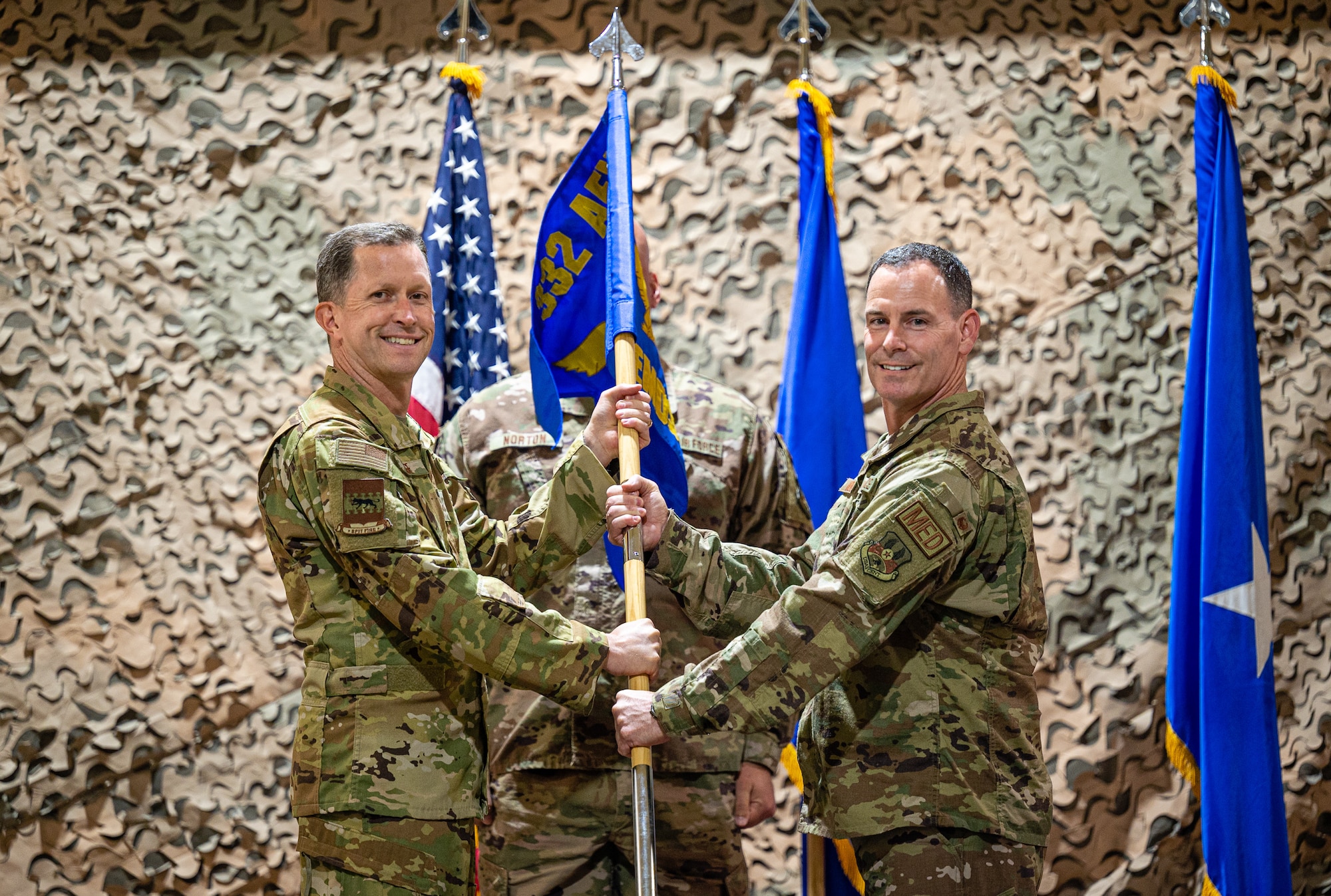 U.S. Air Force Brig. Gen. Christopher Sage, 332d Air Expeditionary Wing commander, exchanges the 332d Expeditionary Medical Squadron guidon with U.S. Air Force Colonel Charles Moniz, 332d EMS incoming commander, during an assumption of command ceremony at an undisclosed location in Southwest Asia, June 24, 2022. The assumption of command is a military tradition that formally signifies the transfer of responsibility from one commanding officer to another, symbolized through the passing of a guidon. (U.S. Air Force photo by Master Sgt. Christopher Parr)