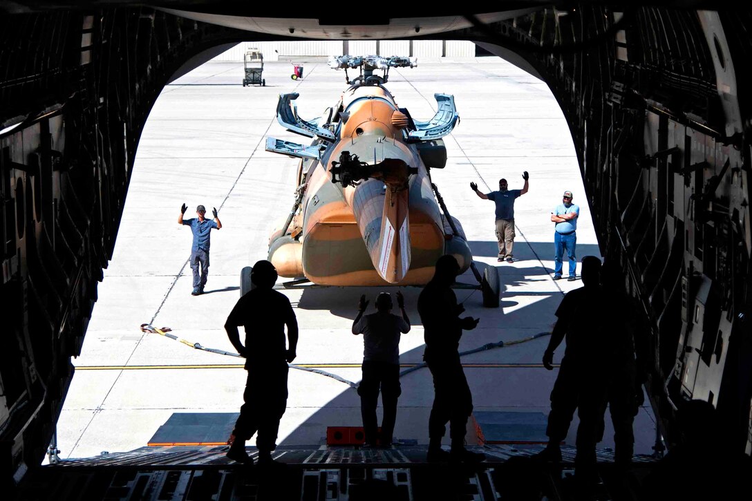 A group of people stand inside and outside the cargo hold of an aircraft preparing to onboard a helicopter.