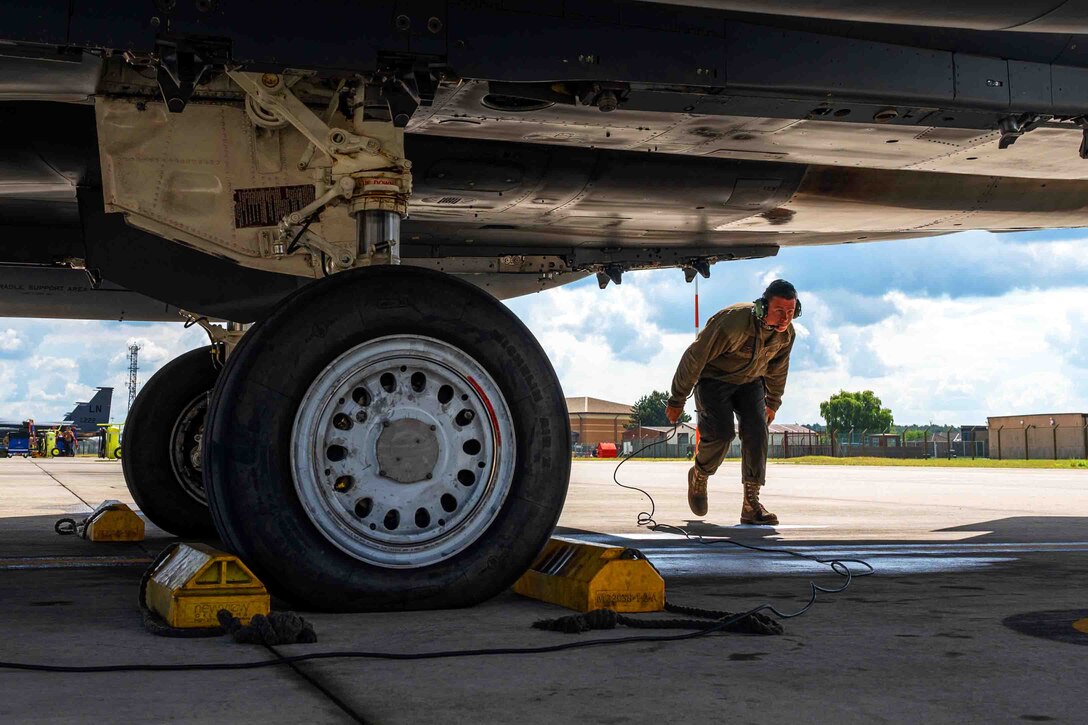 A service member crouches while walking under an aircraft.