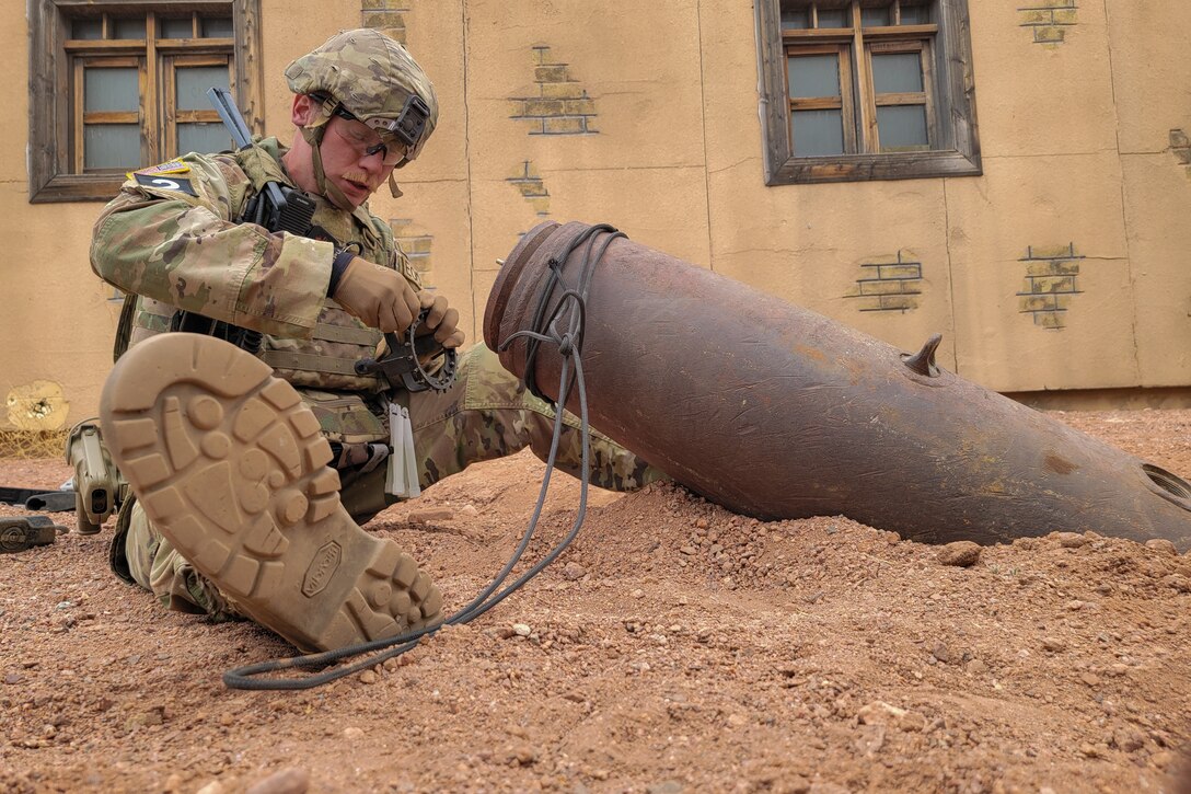 A soldier sits in the dirt and works with a gadget.