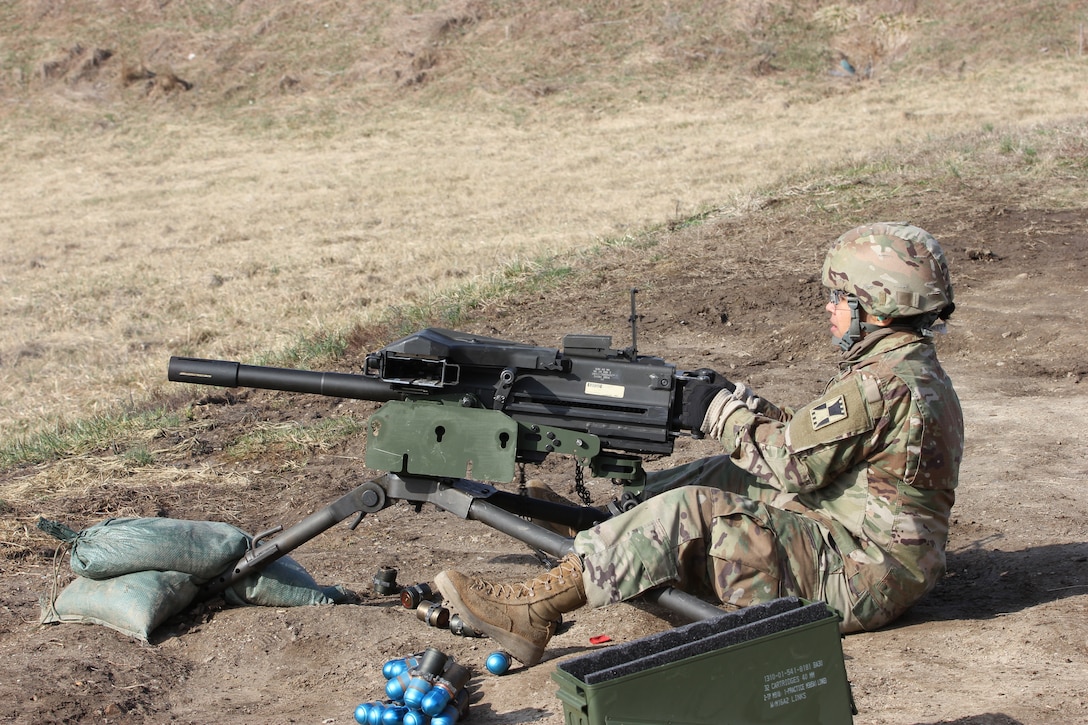 A uniformed service member sits on the ground while operating a military-style weapon.