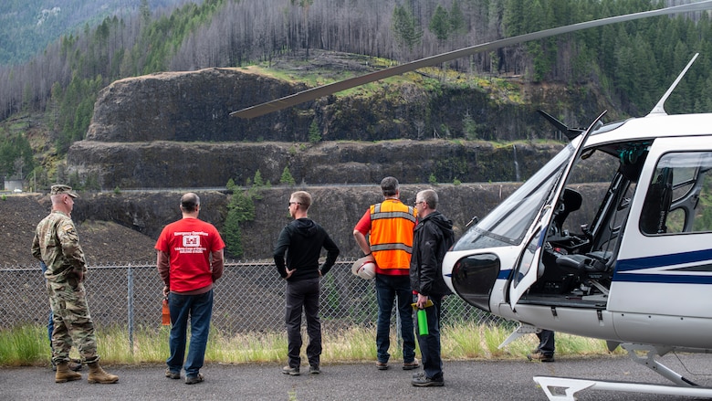 Five men standing in a parking lot next to a helicopter look out over a large, rock-covered dam and river valley.