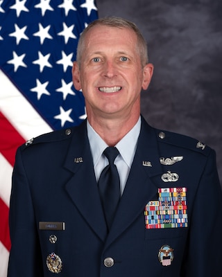Air Force Col. Craig Lowery, a smiling white man with gray hair in a Air Force blue dress uniform with ribbons and badges in front of a US Flag.