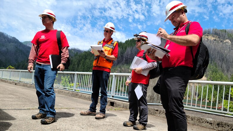 Four men in bright red t-shirts and white protective hard hats write notes in documents on clipboards. It's a mostly sunny day. The men are standing on a road that goes across the top of a dam. Hills dotted with evergreen trees sit off in the distance.