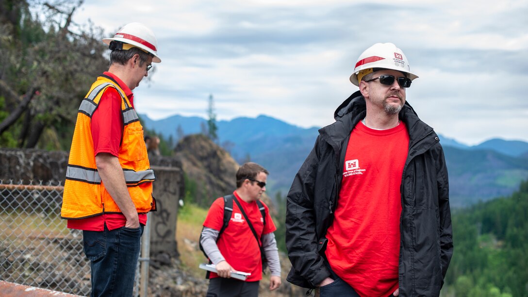 Three men standing on top of a dam, who are wearing bright red t-shirts and white protective hard hats, look down at a river valley covered in evergreen trees.
