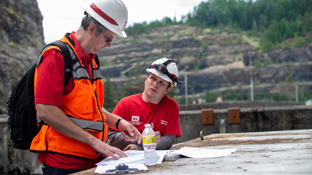 Two men wearing bright red t-shirts and white protective hard hats look at documents on a clipboard on a slightly overcast day. The men are performing a dam safety inspection.