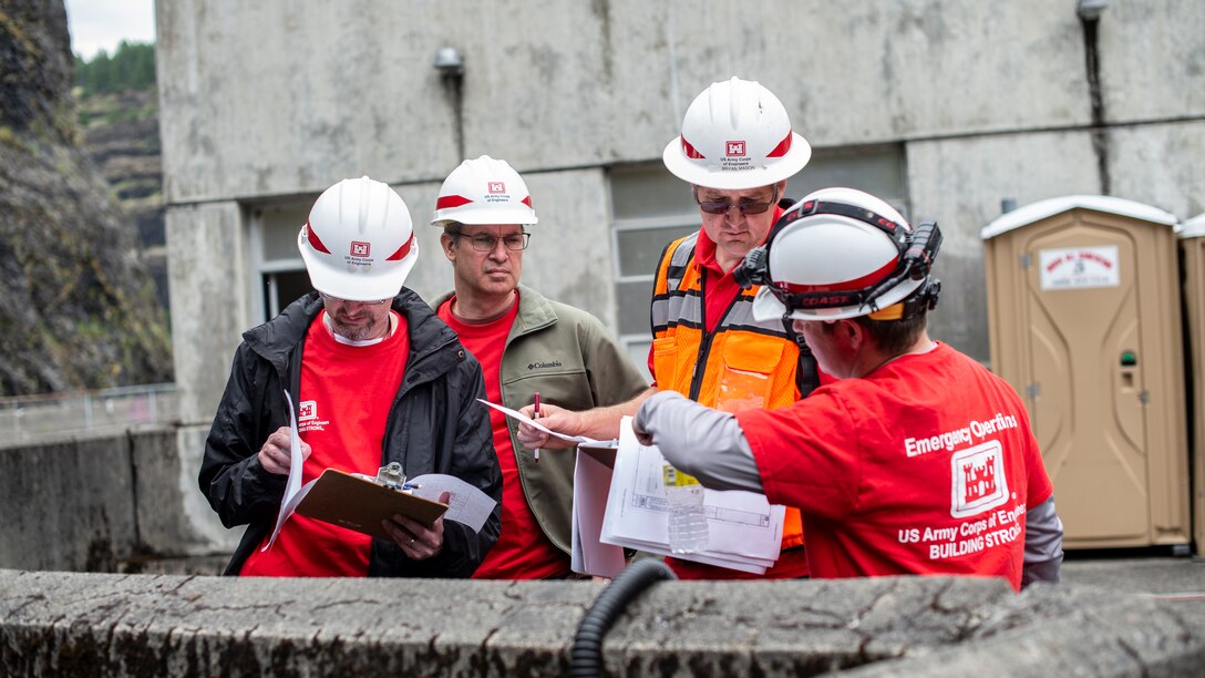Four men wearing bright red t-shirts and white protective hard hats look at documents on a clipboard on a slightly overcast day. The group is performing a dam safety inspection.
