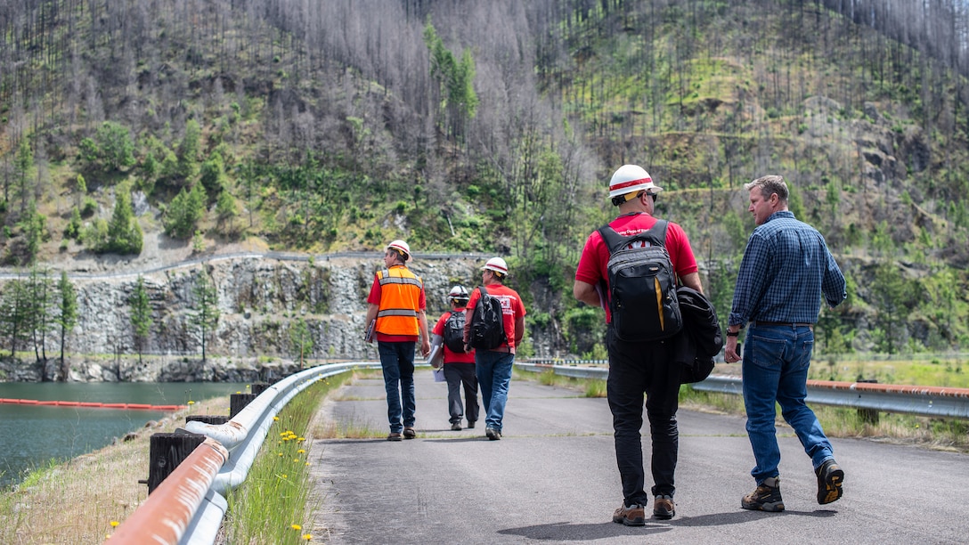 Five men walk down a road, their backs facing the camera, on a sunny day. Hills dotted with evergreen trees sit off in the distance, surrounding a lake. The road goes across the top of a dam.