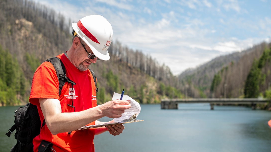 A man in a bright red shirt with a black backpack looks at a form on a clipboard on a sunny day. Hills dotted with evergreen trees sit off in the distance, surrounding a lake.