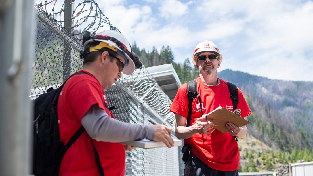 Two men holding clipboards stand near a fence topped with razor wire on top of a dam on a sunny day. Hills dotted with evergreen trees sit off in the distance.