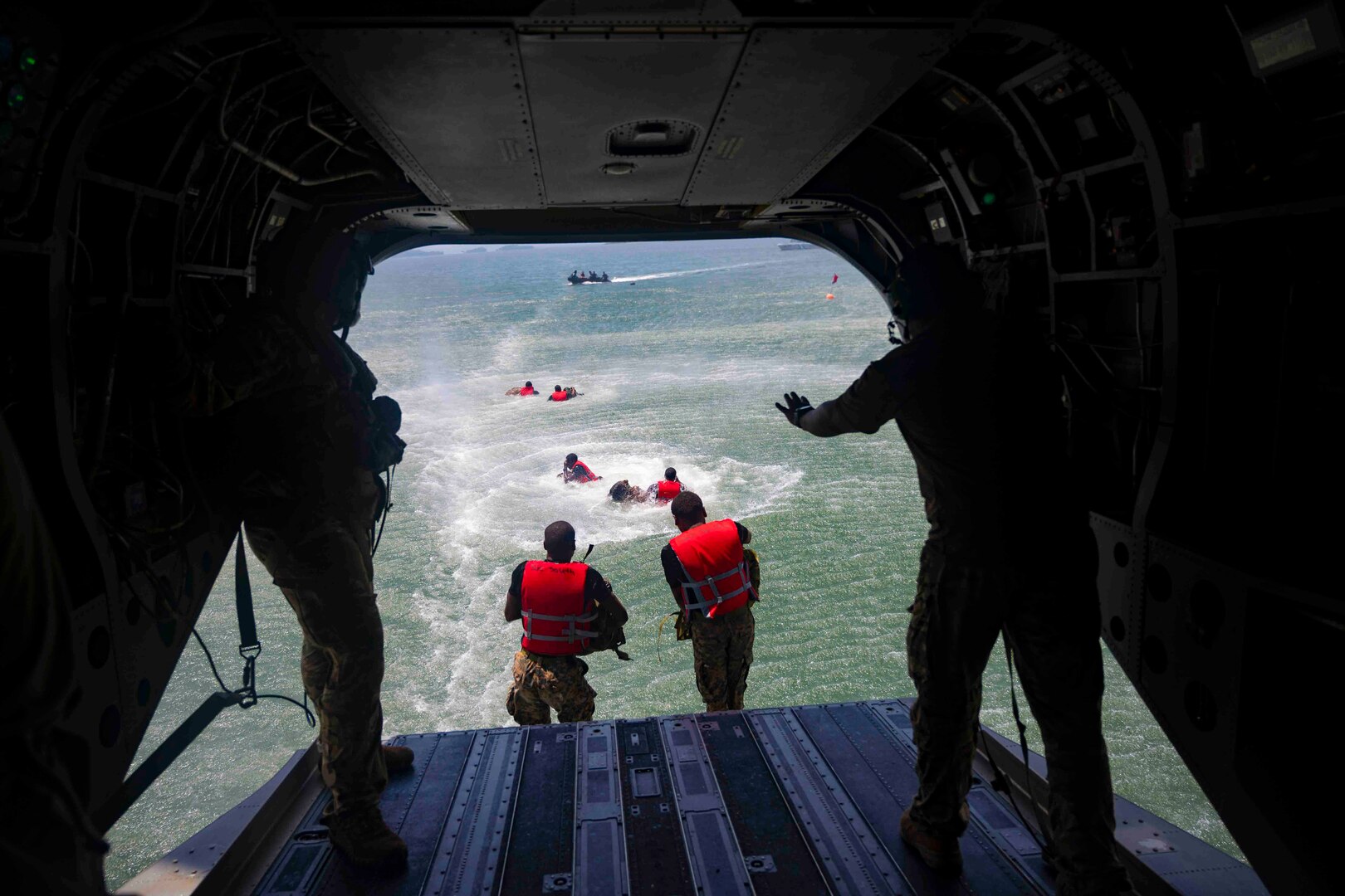 Service members wearing red life vests jump into water from an airborne aircraft.