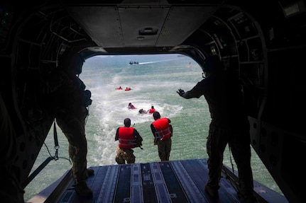 Service members wearing red life vests jump into water from an airborne aircraft.