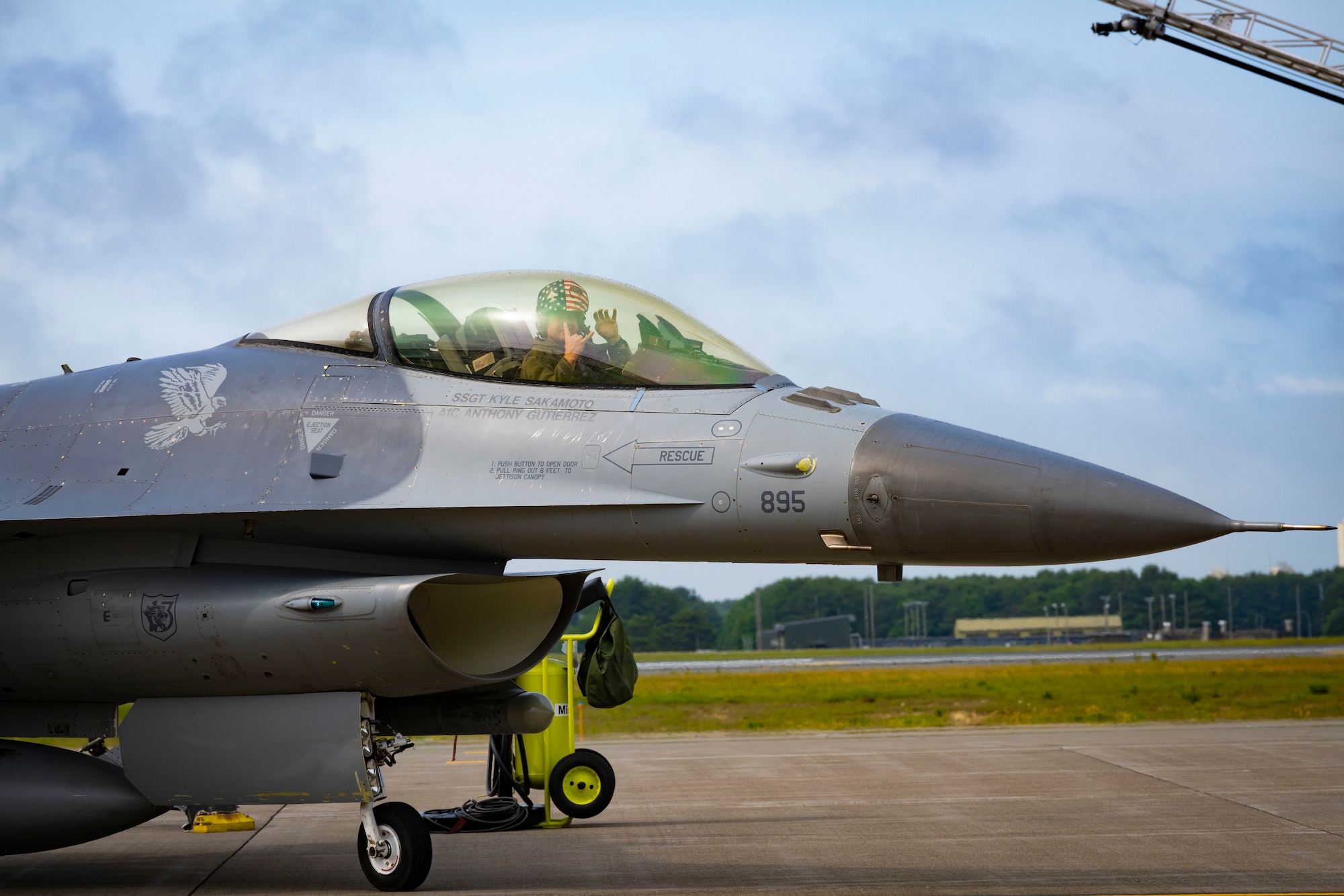 U.S. military member in uniform holds up hand signals while sitting in the cockpit of an F-16 Fighting Falcon.