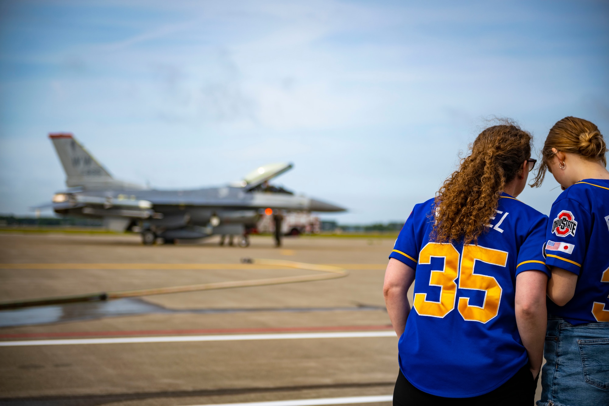 Two U.S. civilians stand on the side of a flight line while an F-16 Fighting Falcon cockpit opens in the background.