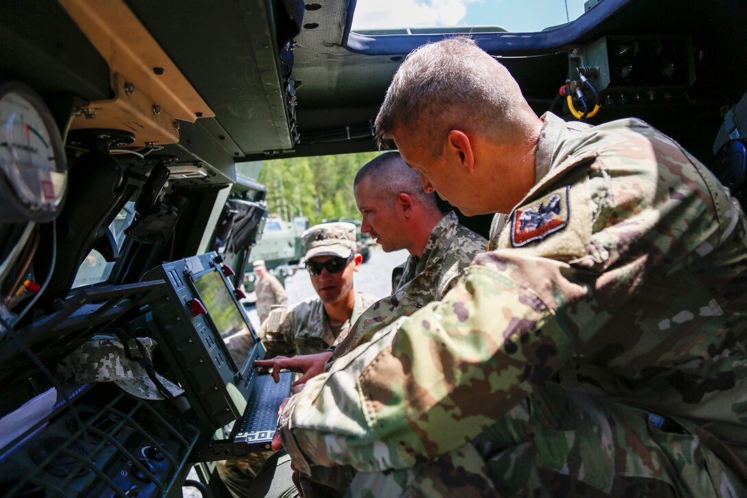 Two service members sit in a military vehicle and interact with an instrument panel while another service member looks in through the window. member looks in through the window