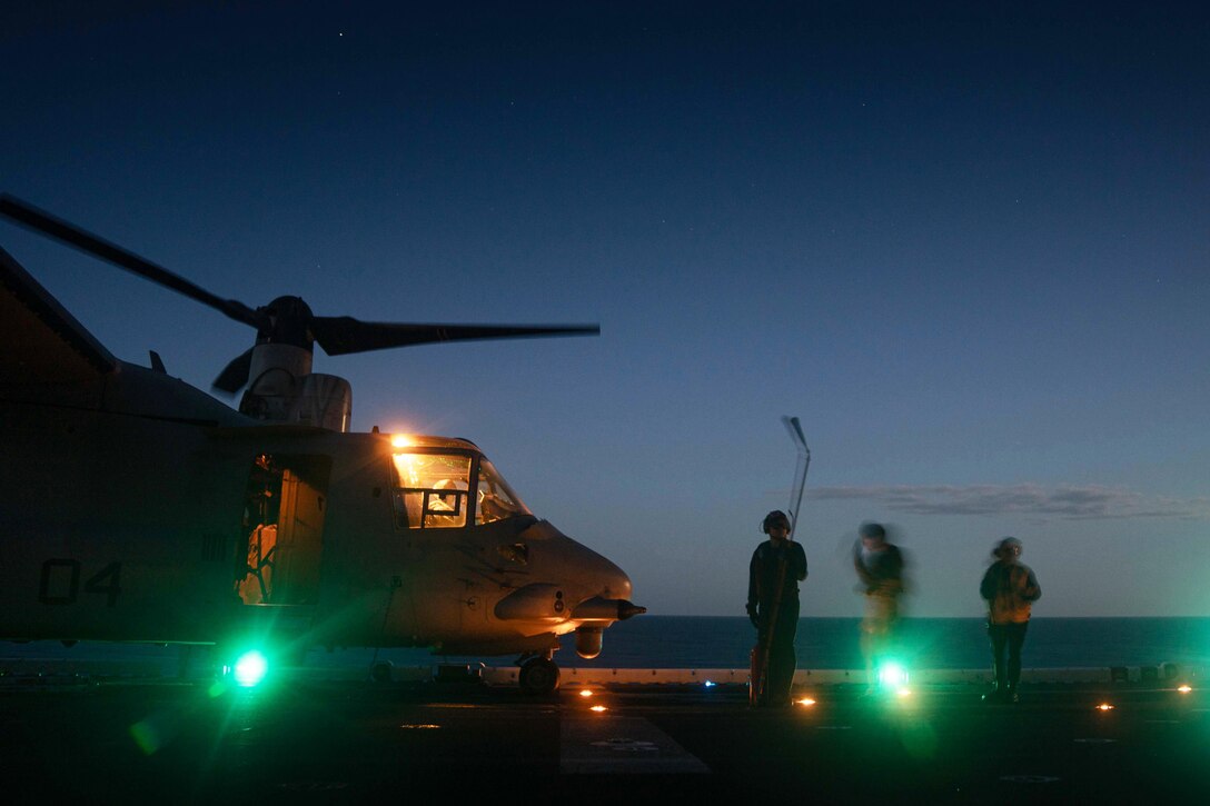 Service members stand on the flight deck next to a military aircraft at night.