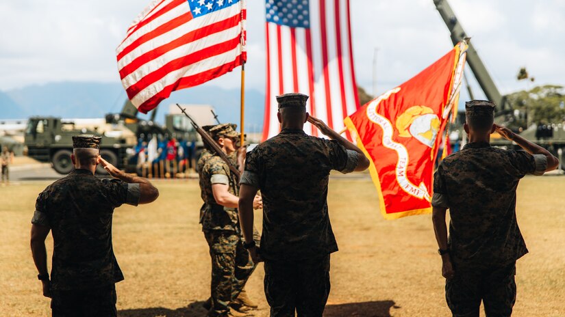 Marines salute as others carrying flags march by them on a field.
