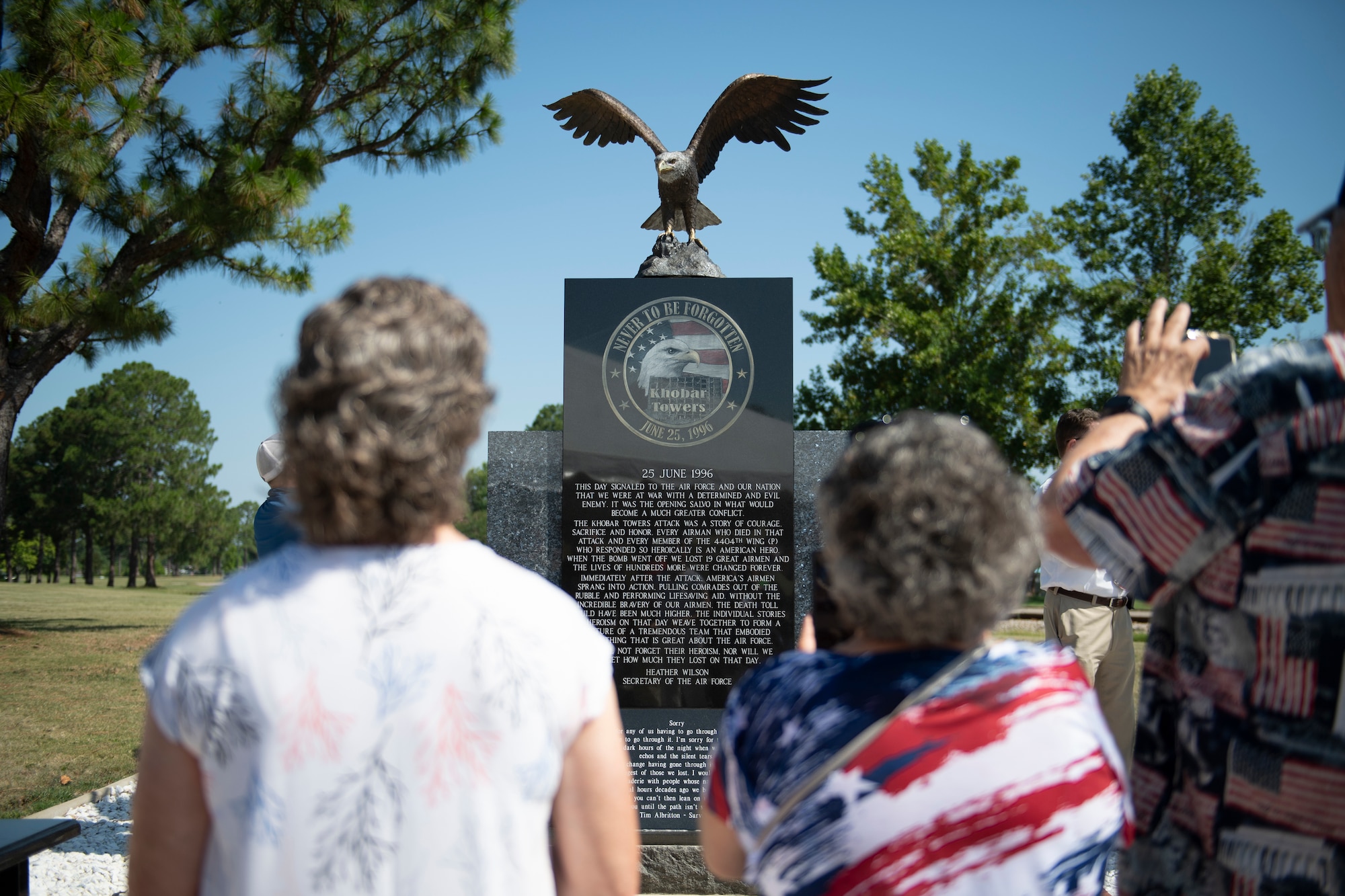 A photo of a monument with an eagle on top of it in the background with three people in the fore ground holding their phones up  to take a picture of the monument.