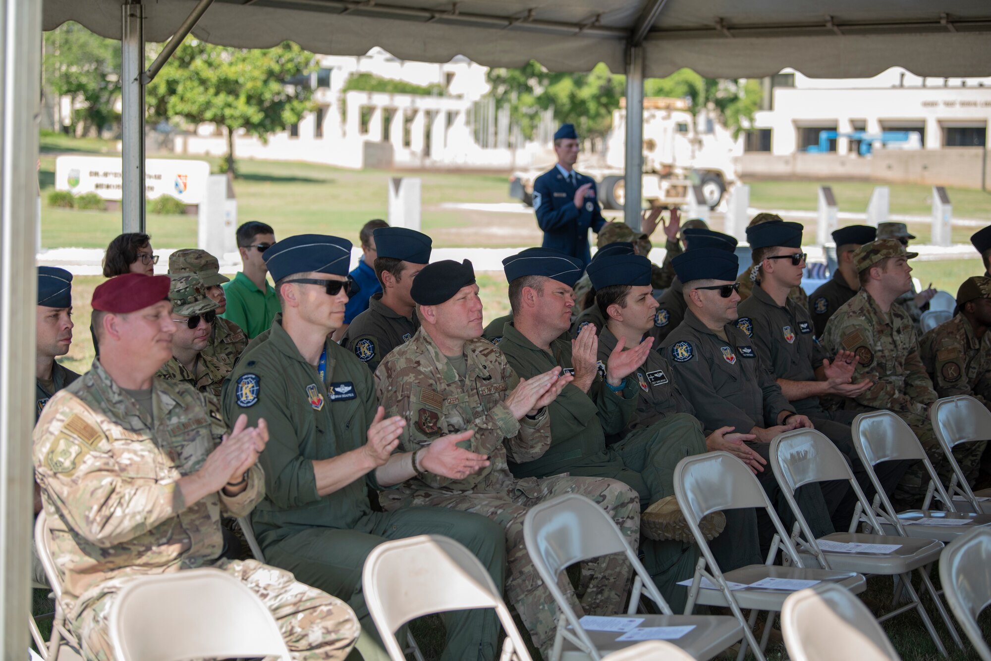 A photo of multiple people sitting in a row of chairs under a tent, clapping.