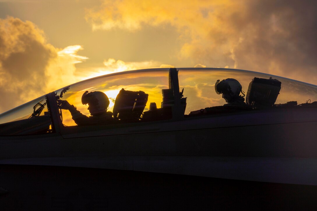 Two Marines sit in an aircraft under a sunlit sky.