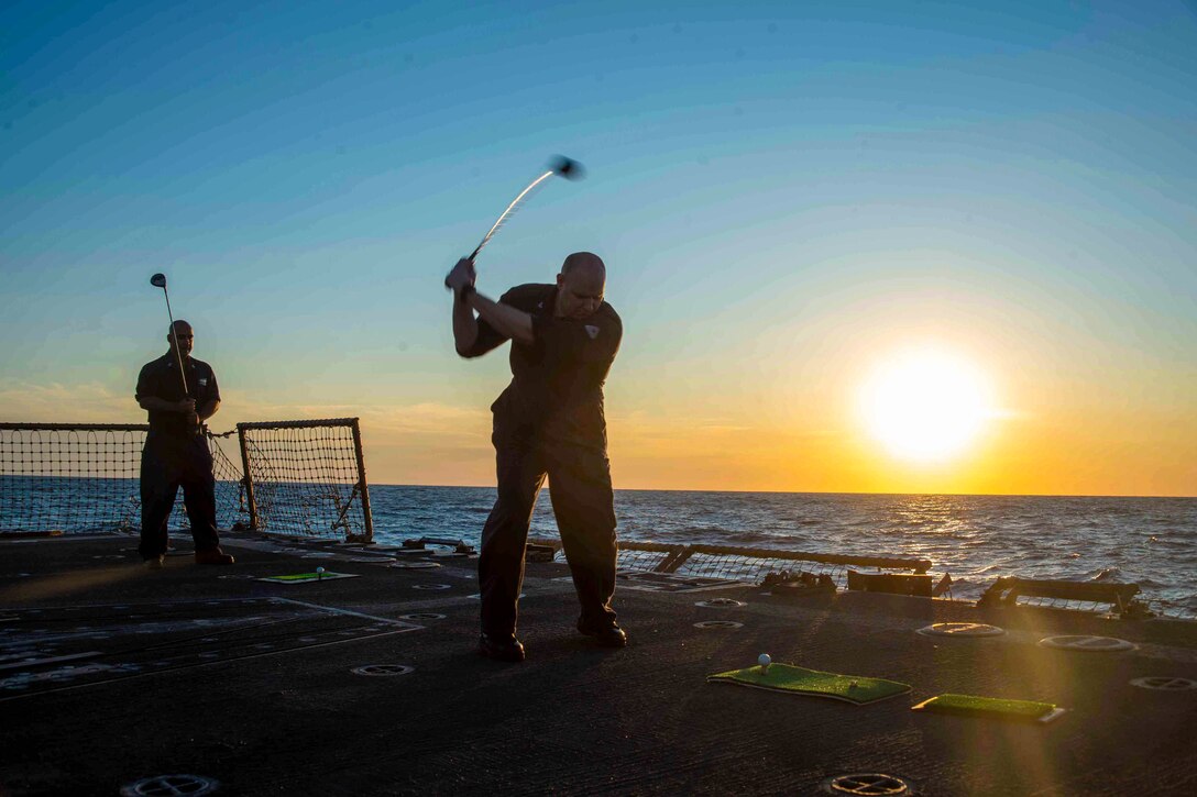 Two sailors hit golf balls with golf clubs on the deck of a ship.