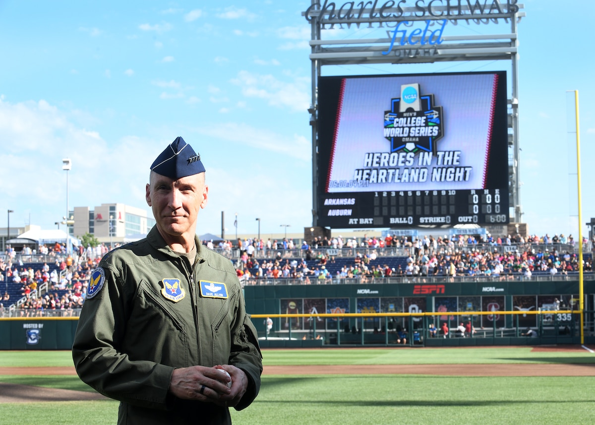 Man in a flight suit stands on a baseball field with a big screen and some of the crowd seen in the background