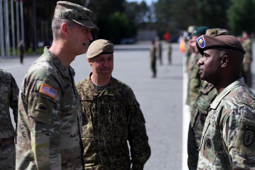A uniformed service member talks to another uniformed service member while a foreign military member looks on.