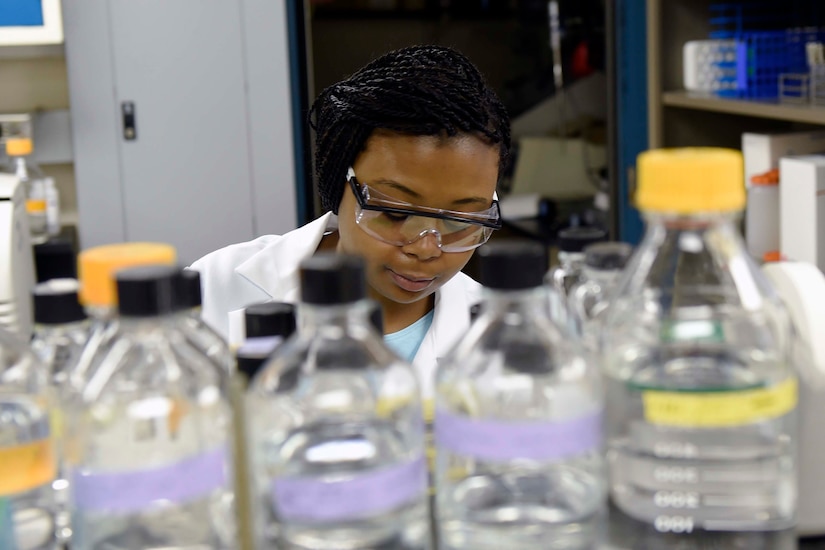 A woman wearing a lab coat and safety glasses stands behind an array of bottles containing liquids.