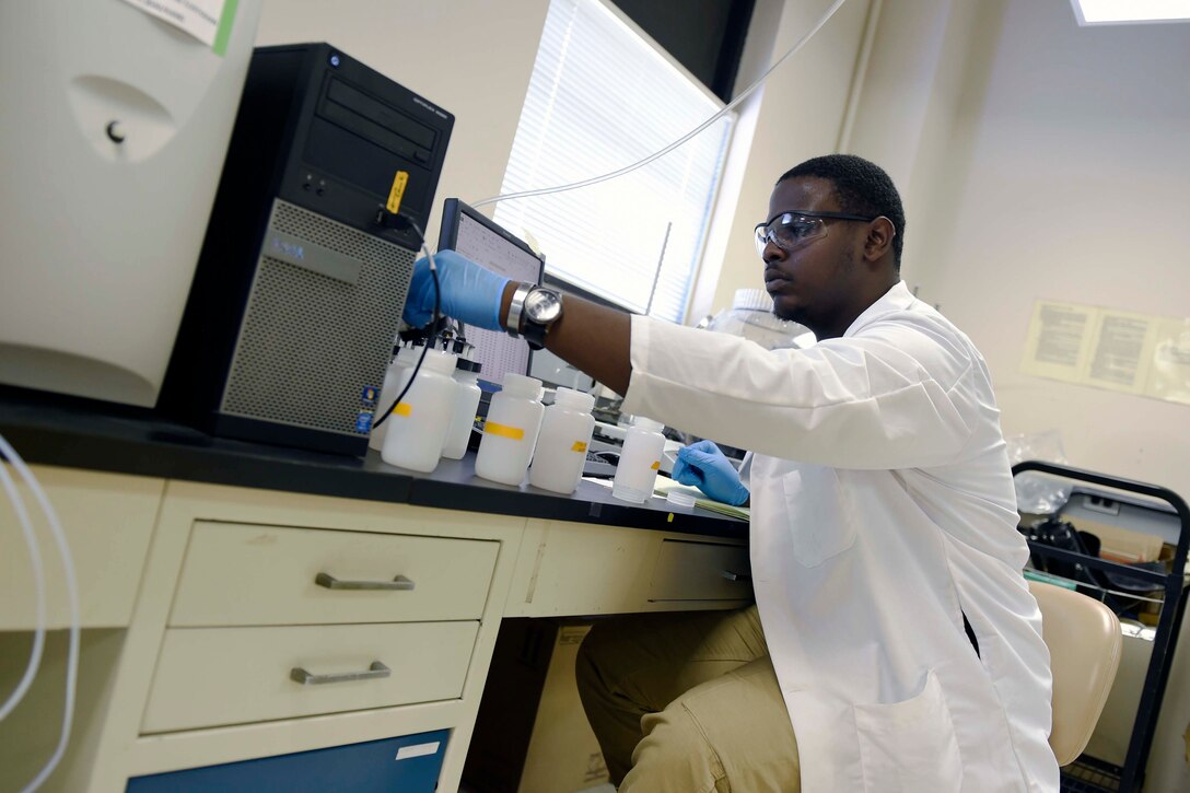 A man wearing a lab coat sits in front of several bottles at his computer desk.