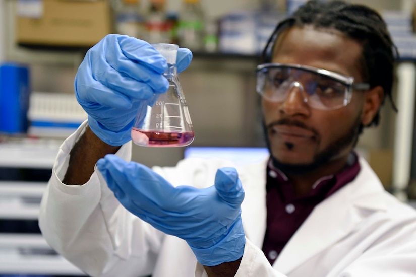 A man wearing a lab coat, safety glasses and latex gloves holds a flask containing a purple liquid.