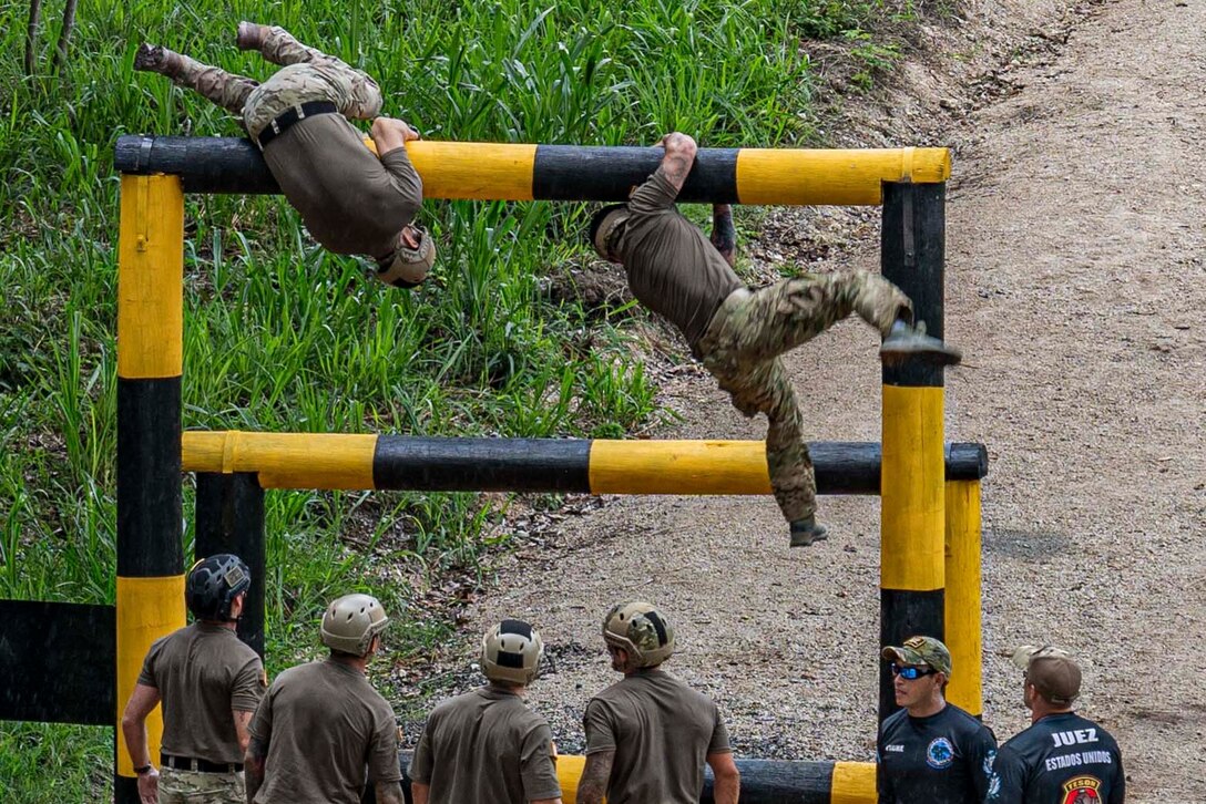 Two soldiers climb over an obstacle as others watch below.