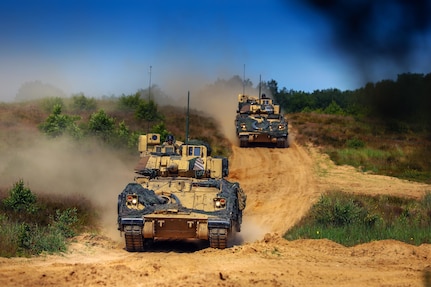 Two armored military vehicles drive down a dirt road and kick up dust in a field.