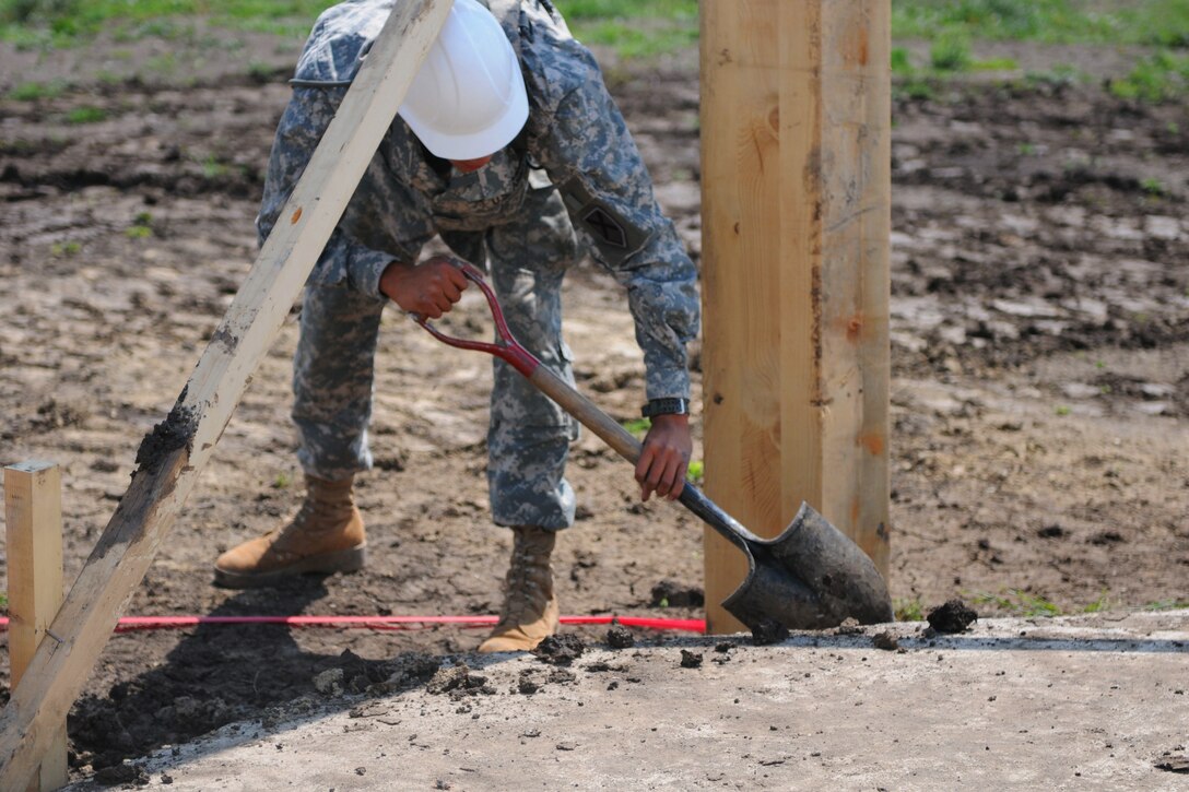 A soldier leans over while holding a shovel and fills a post hole for a multipurpose building