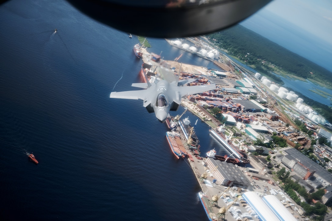 A fighter jet flies at a low altitude over a body of water near a port.
