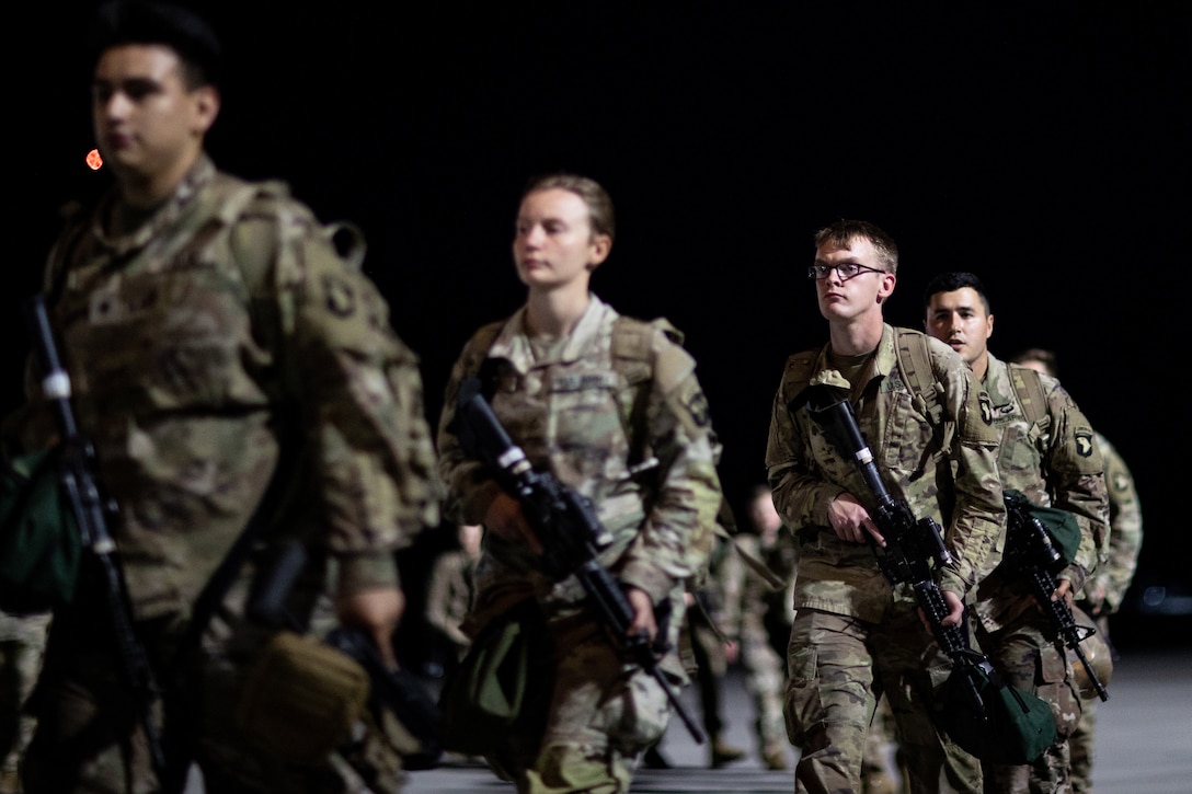 Armed soldiers carrying gear walk in a line.