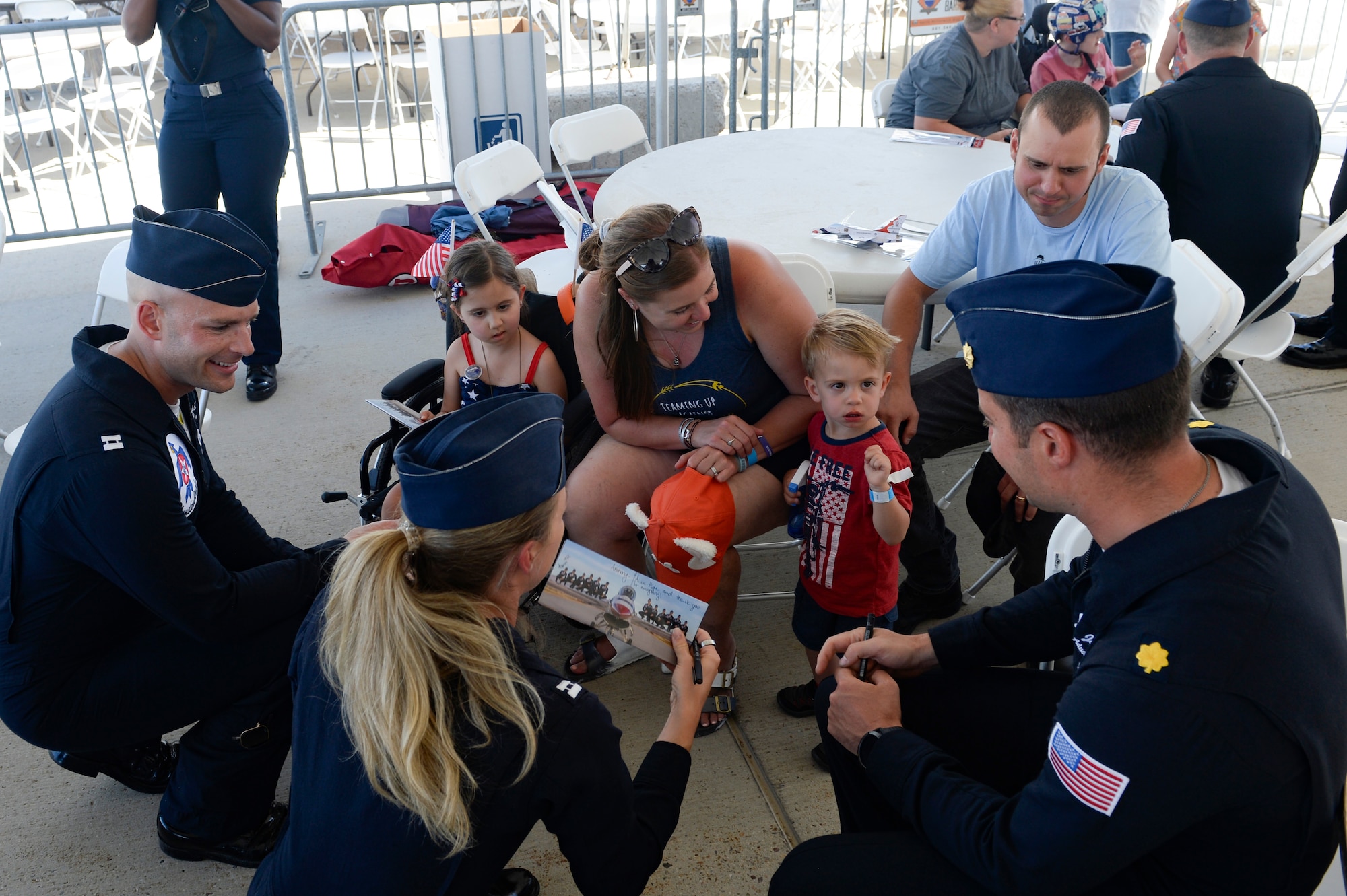 Three Thunderbirds team members kneel down to hand an autographed image to two small children and their parents.