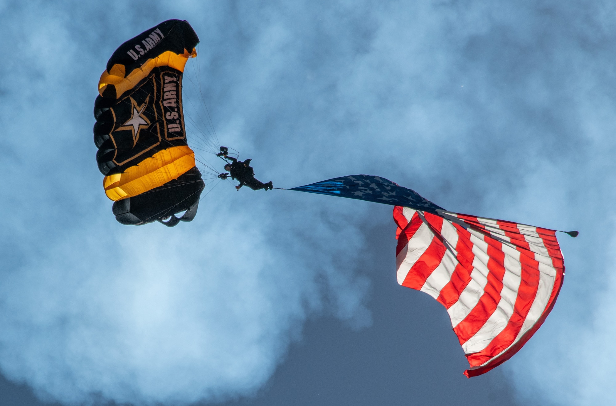 A ground's eye view of a Golden Knight parachuting to the ground. Above him is a black and gold parachute canopy and attached to him is a large American flag.