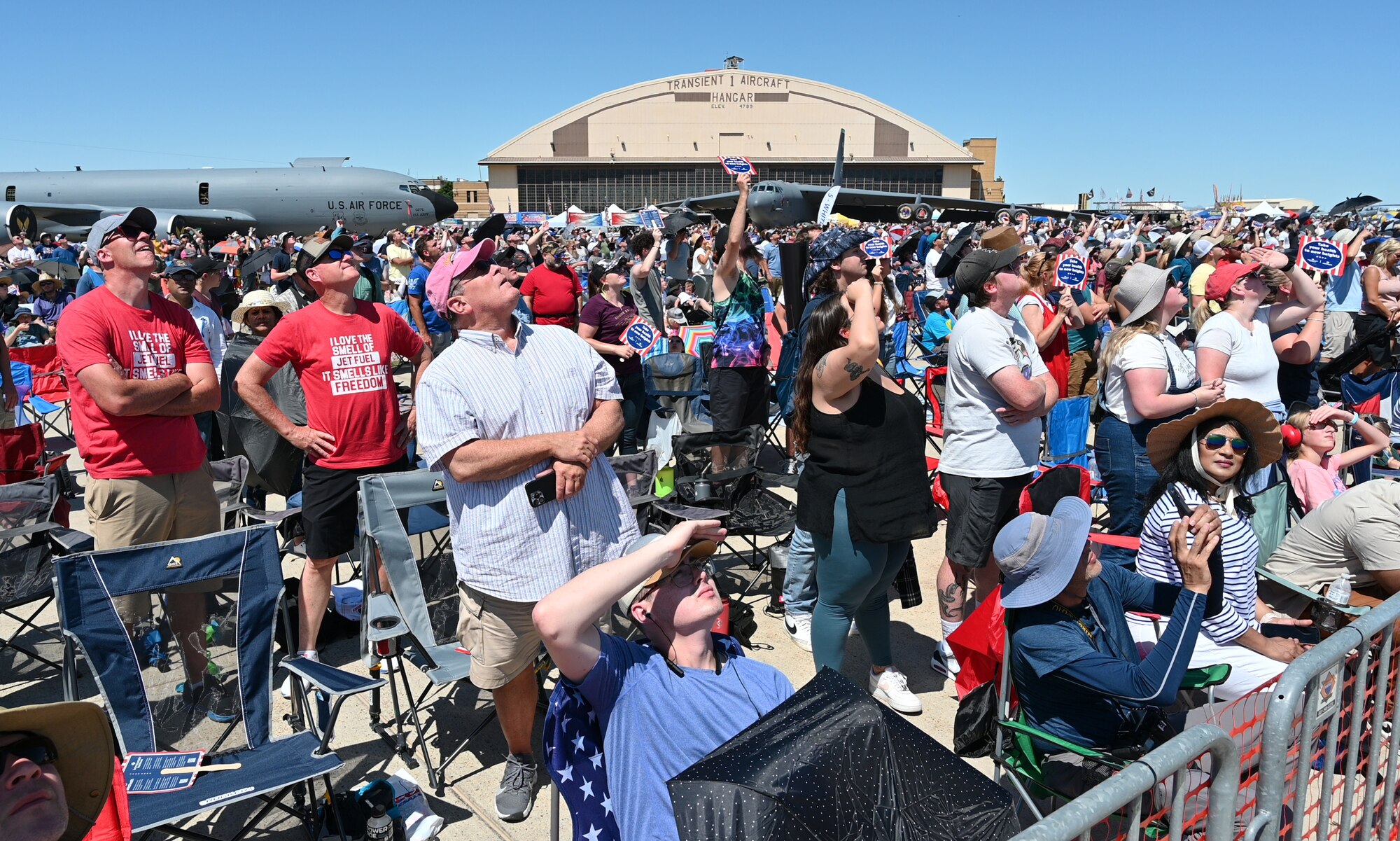 Spectators sit and stand shoulder to shoulder on the airfield looking to the sky watching the air show.