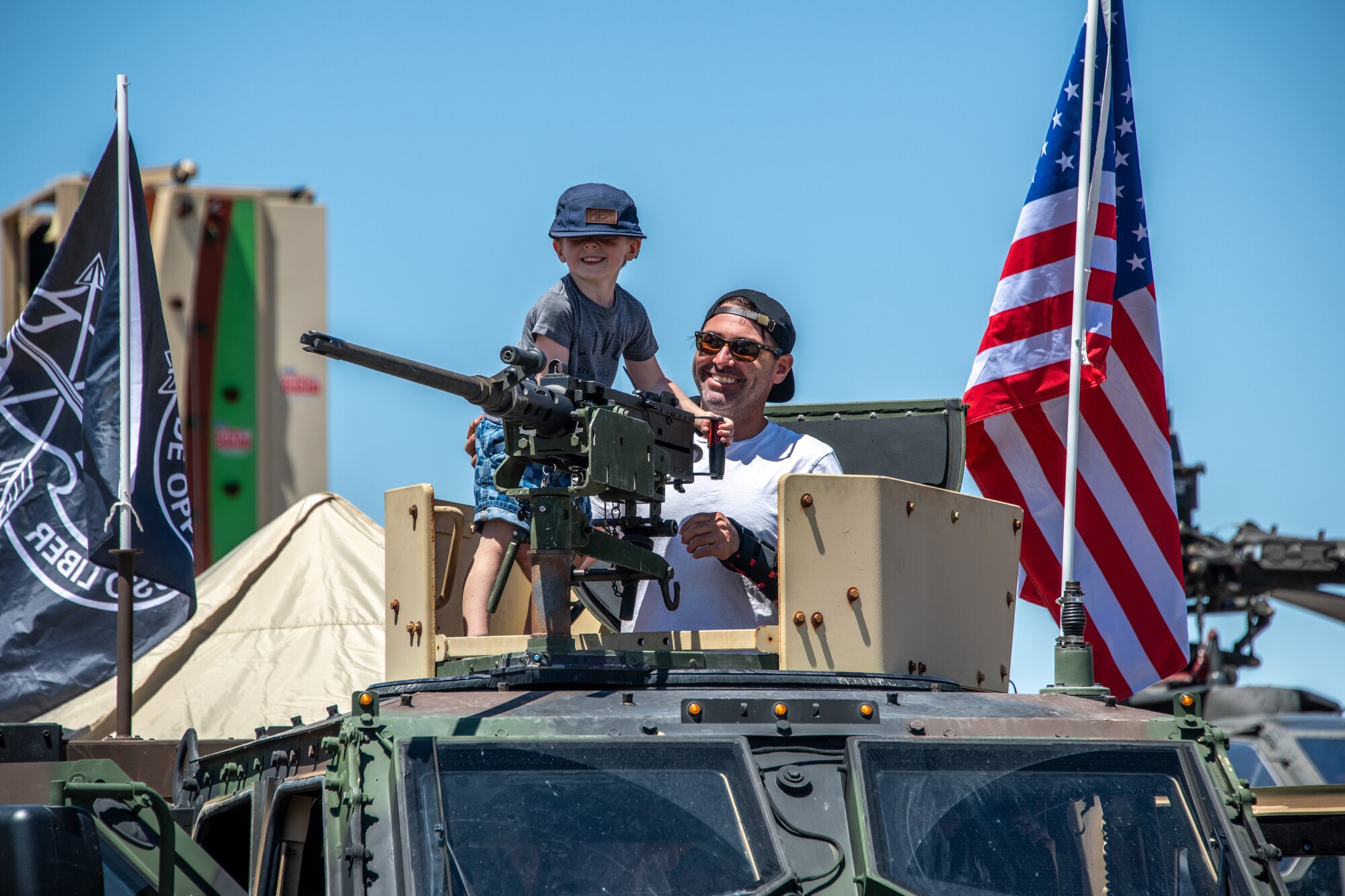 A young boy and man stand inside the turret of a Humvee display with an American flag attached to the vehicle sits on display to the side of them.