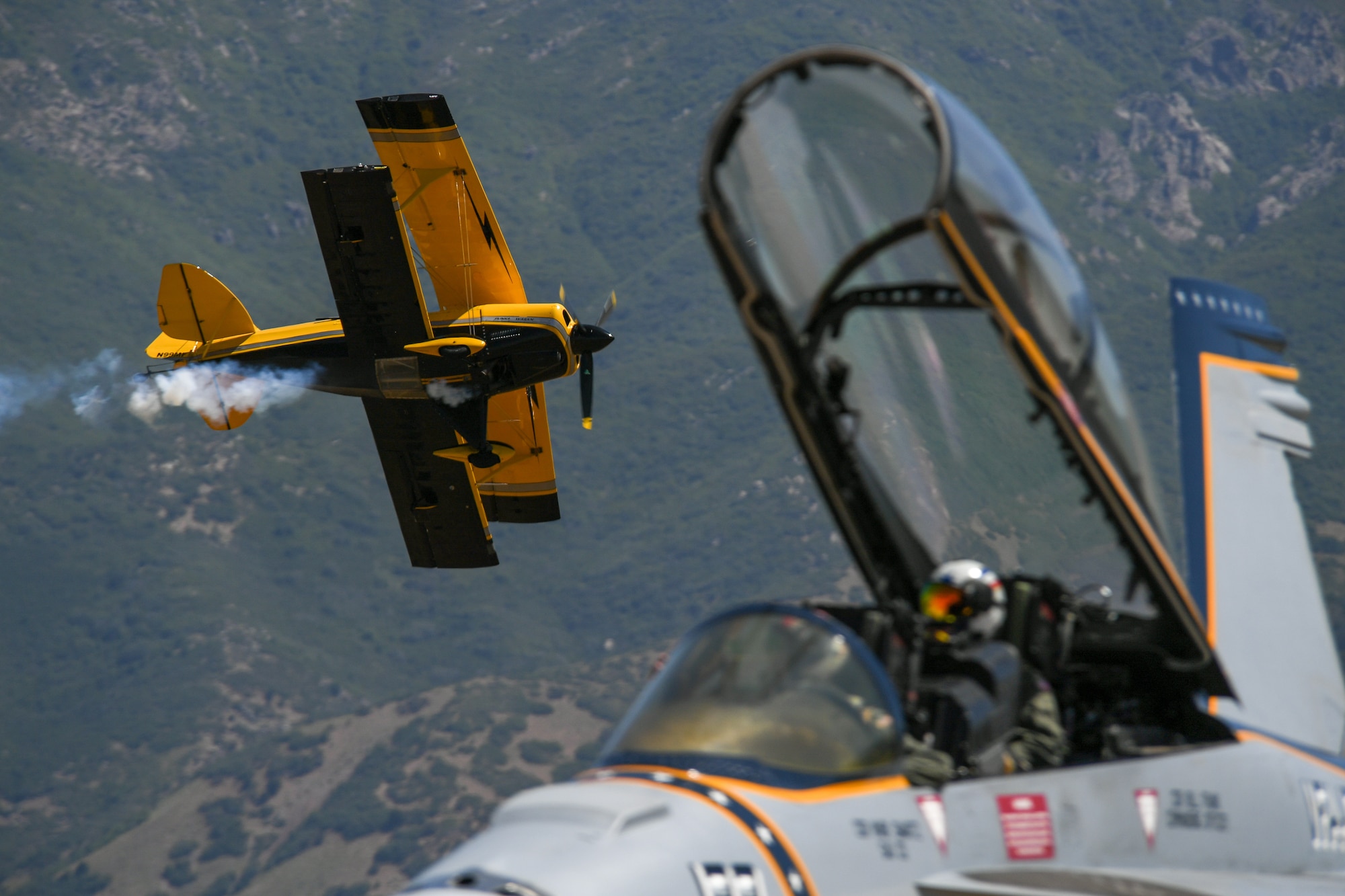 A yellow Pitts S2S flies in the background with while a pilot sits in the cockpit of a U.S. Navy F-18 fighter aircraft in the foreground.