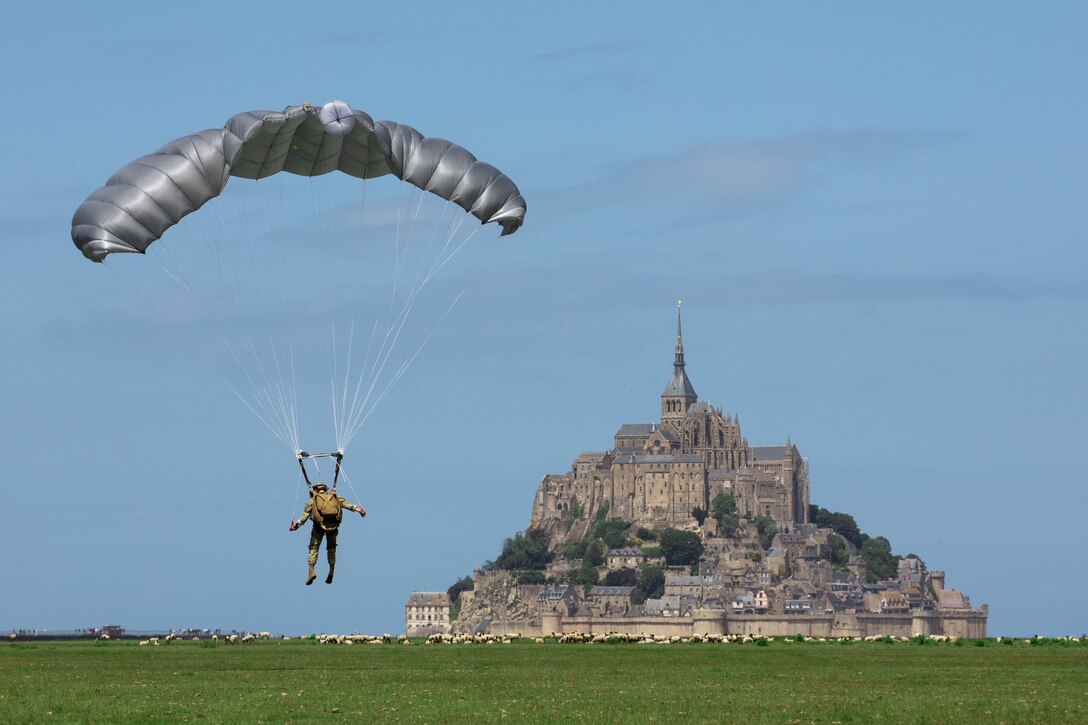 A soldier parachutes to the ground with a large abbey on top of a hill in the background.