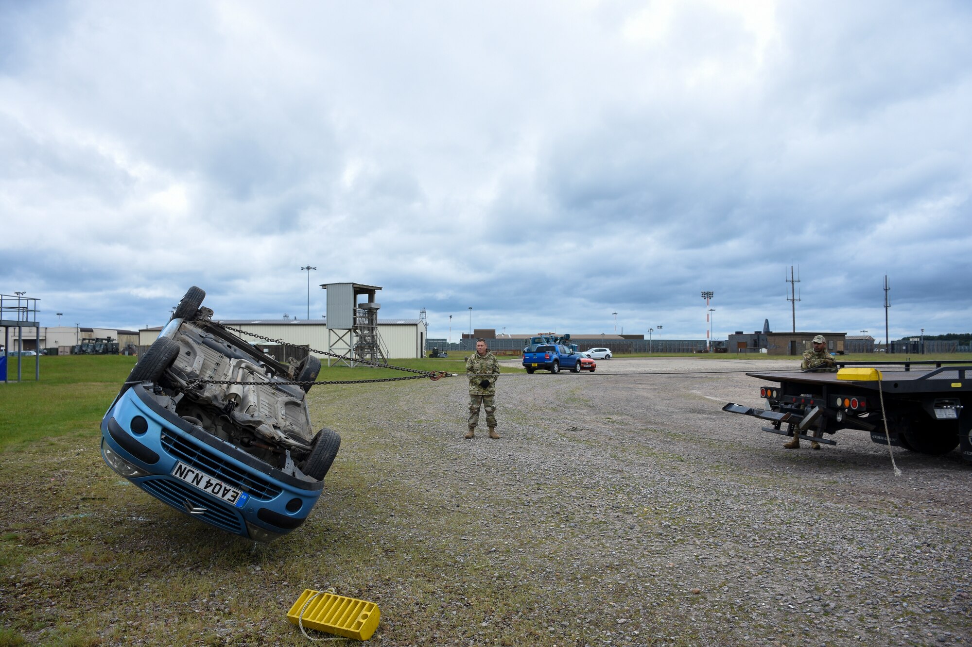 One man operates a winch attached to a car that has been flipped onto its roof while another man observes.