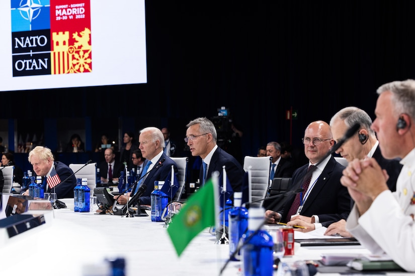 A line of men sit at a long desk with microphones and small national flags placed in front of them.