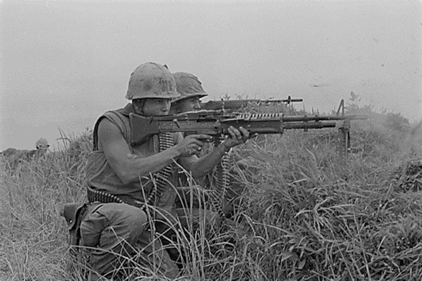 Two men in combat gear aim automatic weapons in tall grass.