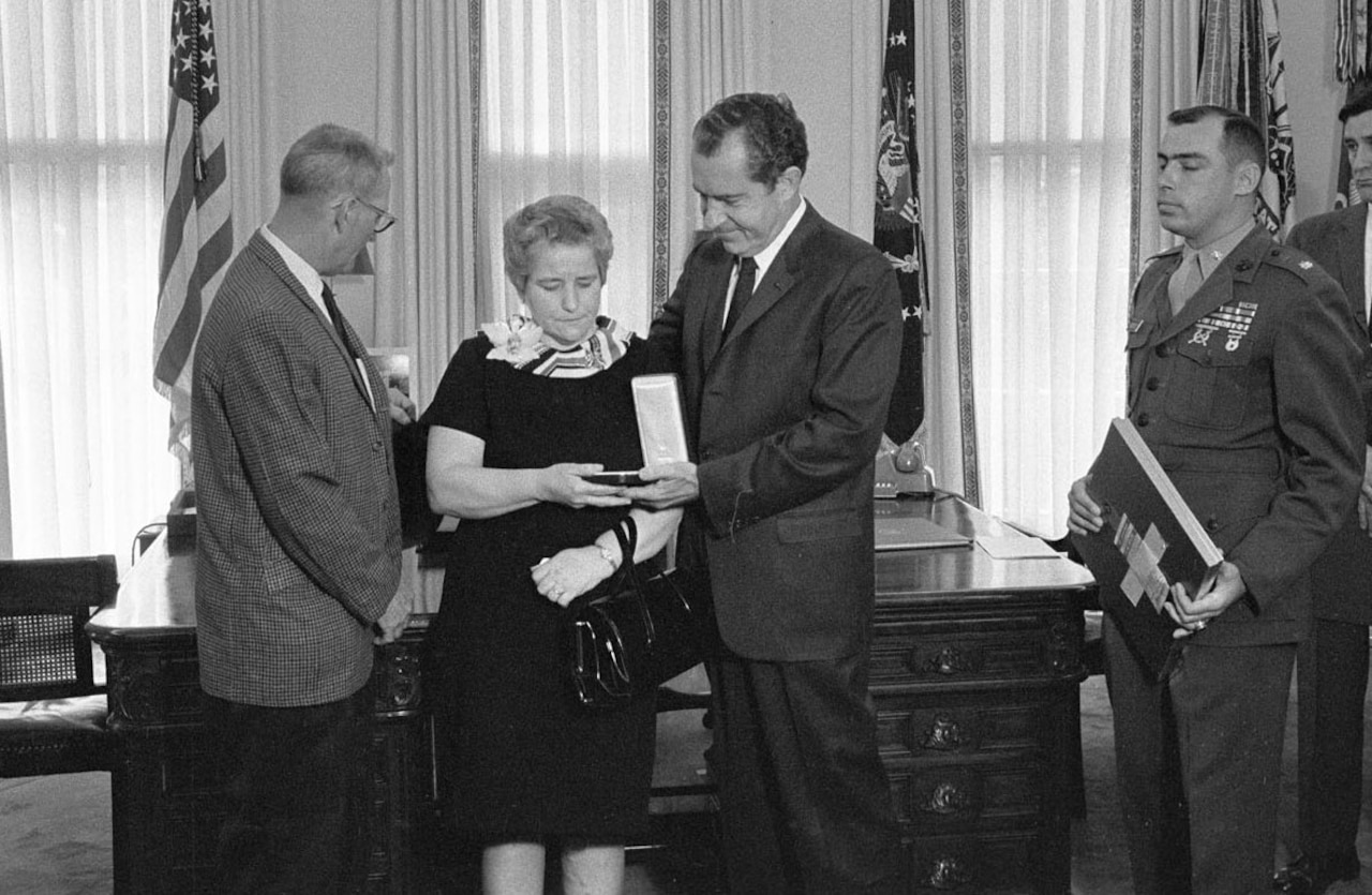 A man presents a medal in a box to a woman. Two men stand nearby.