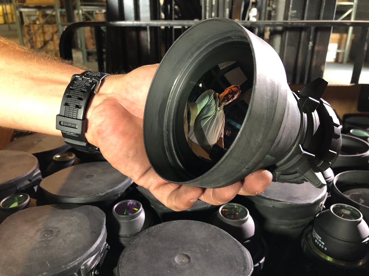 A man holds a camera lens, and his reflection can be seen in its glass.