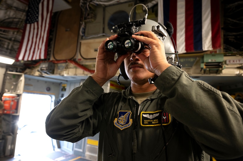 A service member wearing a helmet holds a pair of binoculars up to his eyes and looks through.