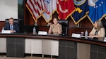 Three people - two women and one man - sit at a table  in front of a row of flags