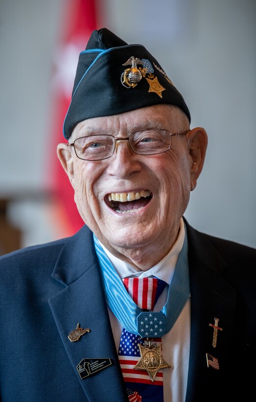Medal of Honor recipient Hershel “Woody” Williams smiles with a flag in the background.